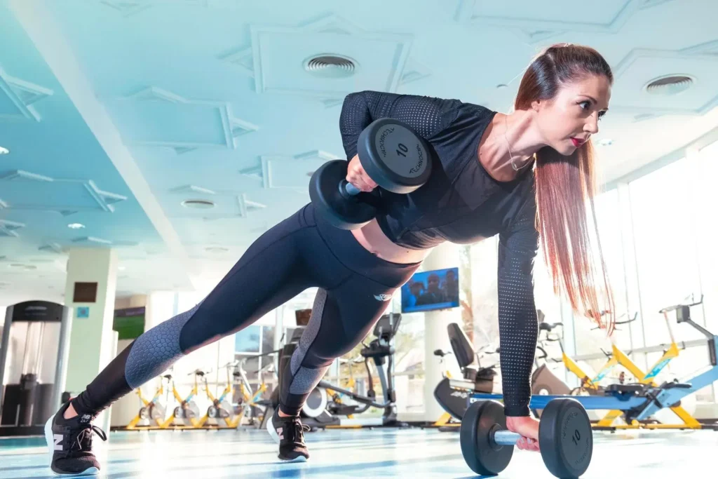 Mulher em treino de academia realizando prancha com halteres, parte de uma rotina de treinos para musculação e plano de treino completo