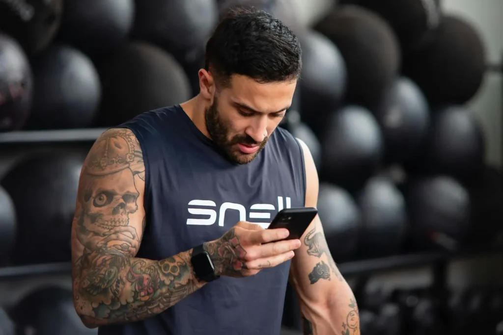 Homem tatuado conferindo cronograma de treino de academia no celular durante treino de musculação